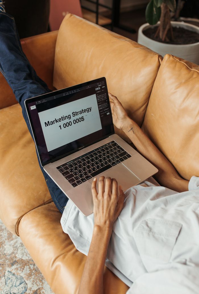 An adult working on a marketing strategy document on a laptop while lying on a couch indoors.